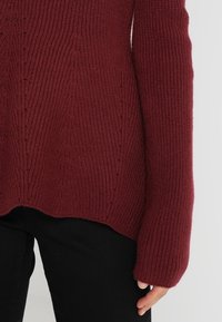 Maglione lavorato a maglia bordeaux con texture a coste indossato con pantaloni neri, maniche che coprono il polso e parte della mano, su sfondo chiaro.