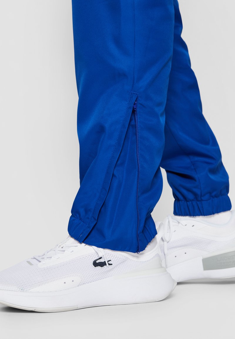 Pantalons de sport bleu roi avec des ouvertures latérales zippées, des poignets élastiques, associés à des baskets blanches avec un dessus en maille et une semelle en caoutchouc.