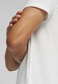 Witte polo shirt van een gestructureerde stof, met een opgerolde mouw en een close-up van een hand die op de arm rust, waarbij een gladde huid zichtbaar is.