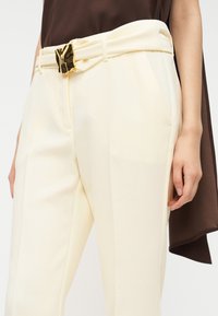 Κρεμ παντελόνι με λεία υφή, που διαθέτει πλαϊνές τσέπες και χρυσή διακοσμητική αγκράφα σε φαρδιά μέση.