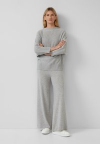 Femme se tenant les bras croisés, portant un pull gris ample, un pantalon large gris et des baskets blanches, devant un fond clair et uni.