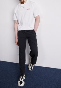 Man som bär en vit Timberland-t-shirt, svarta cargo-byxor och svart-vita sneakers, står på en blåmönstrad matta mot en vit vägg.