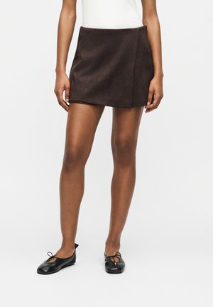 Femme portant une mini-jupe en daim brun foncé et des ballerines noires, debout sur un fond blanc uni.