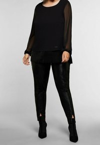 Blusa nera a maniche lunghe in tessuto trasparente con orlo decorato, abbinata a pantaloni in pelle nera lucida e stivali con tacco e cinturino alla caviglia.