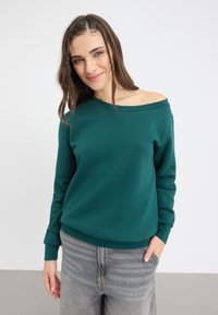 Sudadera verde de hombros al descubierto en material suave, con mangas largas y puños acanalados, combinada con vaqueros de tiro alto grises.