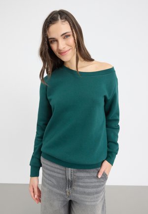 Grønn av-skulderen sweatshirt i mykt materiale med lange ermer og ribbestrikkede mansjetter, kombinert med grå high-waisted jeans.