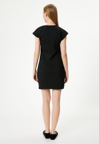 Schwarzes kurzes Kleid mit figurbetontem Schnitt, gefertigt aus glattem Material und mit einem klaren Ausschnitt. Das Model trägt elegante flache Schuhe mit einem dezenten Schnallen-Detail.