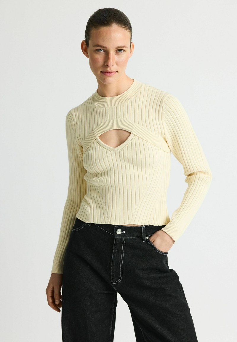 Pull en crème côtelé avec une découpe au niveau du décolleté et des manches longues, associé à un jean en denim noir taille haute.