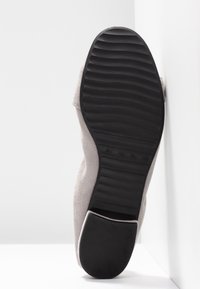 Semelle d'une chaussure en daim gris avec une bande de roulement en caoutchouc noir, posée verticalement contre un angle où se rejoignent un mur blanc et le sol.