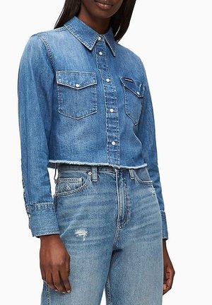 Kobieta nosząca krótką niebieską koszulę jeansową na guziki z klapkowymi kieszeniami oraz niebieskie, wysokowalne jeansy z lekkimi przetarciami.