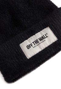 Svart stickad mössa med en mjuk, texturerad yta. Har en sytt vit etikett med svart text som läser "OFF THE WALL" och skötselråd.