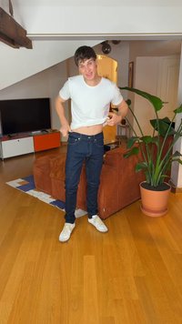 Egy személy fehér pólót és sötétkék farmert visel, és éppen igazítja a farmert. A háttérben egy kanapé és egy cserepes növény látható.