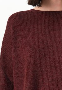 Gros plan sur une personne portant un pull côtelé en maroon avec un col rond et des cheveux bruns courts.