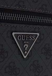 Matériau noir texturé avec un logo triangulaire argenté portant l'inscription "GUESS USA." Comprend une fermeture éclair avec des coutures visibles.