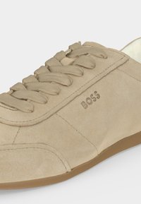 Baskets basses en daim beige avec lacets assortis, semelle en caoutchouc marron et logo "BOSS" embossé sur le côté.