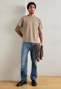 Beige pamut póló rövid ujjakkal és címkével, kék farmernadrággal párosítva. Barna csíkos pulóver a kézben, fekete cipő.