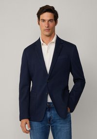 Marineblazer mit glatter Textur, Reverskragen, zwei Vordertaschen, einer Knopfverschluss und kombiniert mit einem weißen Poloshirt und Jeans.