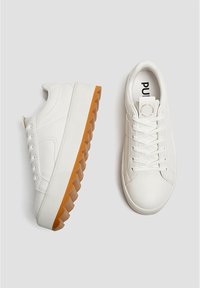 Baskets blanches avec une semelle en caoutchouc texturée, présentant un motif en zigzag en relief. Tige en matériau lisse, fermeture à lacets, et marque sur la semelle intérieure.