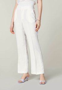 Bílé lněné kalhoty s širokým střihem, které mají dvě přední kapsy a jemnou texturu, doplněné levandulovými sandály na podpatku.