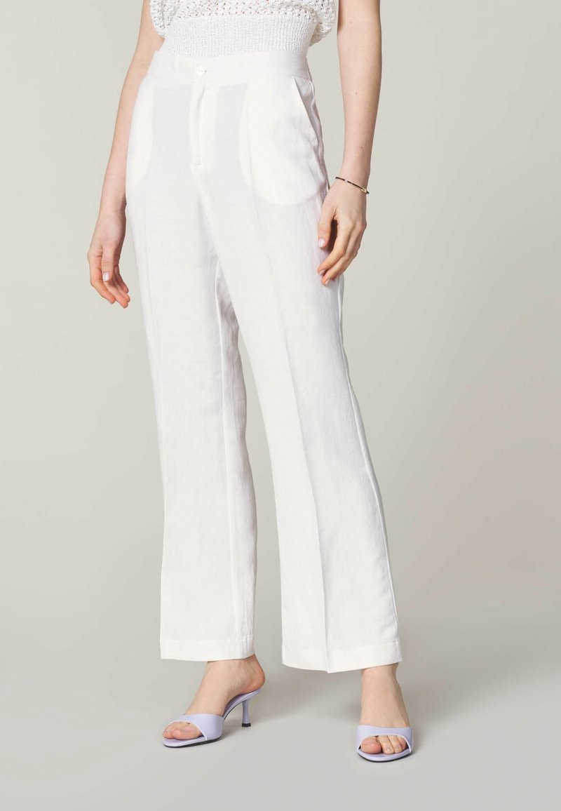 Bílé lněné kalhoty s širokým střihem, které mají dvě přední kapsy a jemnou texturu, doplněné levandulovými sandály na podpatku.
