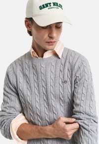 Grå kabelstrik sweater med rund halsudskæring, med et lille broderet emblem. Underneath er en lyse orange og hvid stribet skjorte synlig.