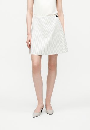 Woman wearing a white pinstriped A-line skirt with a black button and beige pointed-toe heels, standing against a plain white background.