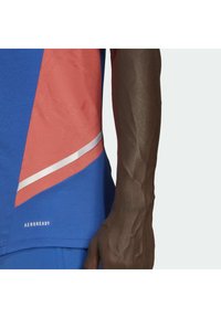 Modré a korálové sportovní tričko s texturovanou tkaninou, s stříbrným pruhem a logem "AEROREADY" na lemu. Detailní záběr ruky.