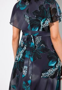 Blumenkleid in dunklen Farben mit türkisfarbenen und weißen Mustern. Hat ein fließendes Design, kurze Ärmel und einen gebundenen Taillenausschnitt. Weiche Textur.