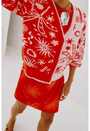 Osoba ubrana w czerwony i różowy kardigan zapinany na guziki z białymi wzorami niebiańskimi oraz czerwoną spódnicę z motywami słonecznymi, a także naszyjnik z zawieszką w kształcie twarzy.