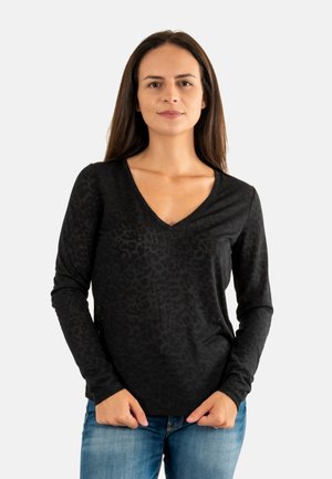 Femme aux longs cheveux bruns portant un t-shirt noir à manches longues et décolleté en V avec un motif léopard subtil et un jean bleu, se tenant contre un fond blanc.