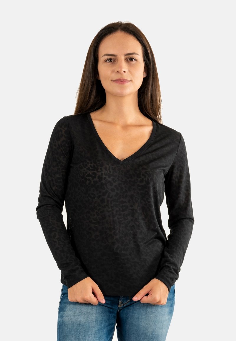 Donna con lunghi capelli castani che indossa una maglietta nera a maniche lunghe con scollo a V e un sottile motivo leopardato, insieme a jeans blu, in piedi su uno sfondo bianco.