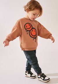 Brązowa oversize'owa bluza z dwoma pomarańczowymi uśmiechniętymi buźkami; ciemnozielone spodnie cargo; czarno-białe sneakersy z rzepami; casualowy strój.