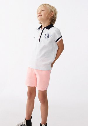 Camiseta polo blanca con cuello azul marino y acentos rosas, con un estampado gráfico; combinada con pantalones cortos rosas y botas negras. Tela suave, corte casual.