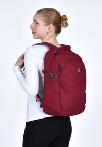 Zaino bordeaux con un design elegante, spallacci imbottiti, diverse tasche con zipper e chiusure laterali regolabili, realizzato in tessuto resistente.