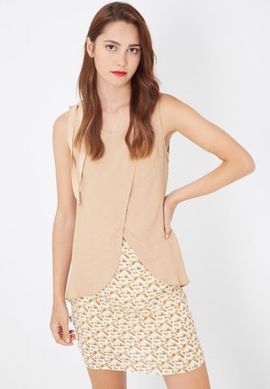 Top sin mangas beige con detalles de cintas, diseño en capas. Combinado con una mini falda blanca con un patrón abstracto colorido.