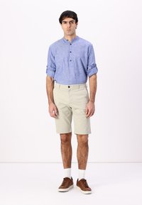Lys blå lin-skjorte med knappeopplæring, kombinert med beige shorts. Brune joggesko og hvite sokker fullfører antrekket. Enkel bakgrunn.