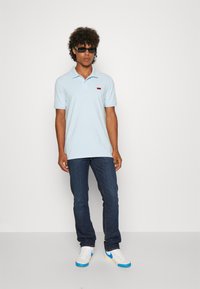 Ljusblå polo-tröja med kort krage, med en liten röd logo på bröstet; i kombination med mörka jeans och vita sneakers med blå accenter.