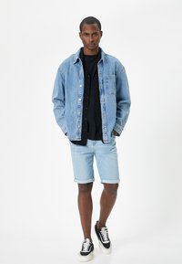 Lysblå denimjakke med forlommer, over en sort skjorte. Lysblå rullede shorts og sorte sneakers med hvide detaljer.