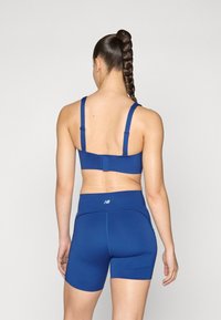 Frau mit geflochtenem Haar trägt einen blauen Sport-BH und passende hoch taillierte sportive Shorts und steht mit dem Rücken zur Kamera vor einem schlichten Hintergrund.