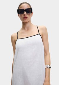 Robe blanche sans manches avec des fines bretelles noires, comportant un tissu texturé. Modèle portant des lunettes de soleil oversize noires et un bracelet en argent.