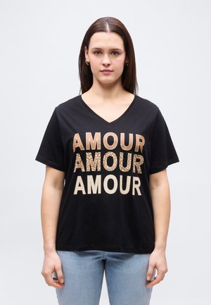 Kvinde med lige brunt hår bærer en sort V-hals t-shirt med "AMOUR" i tre forskellige beige og mønstrede stilarter, kombineret med lyseblå jeans.