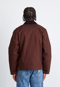 Brown canvas jacket with a dark collar, wide shoulders, and snap closures at the bottom, paired with blue camouflage jeans.