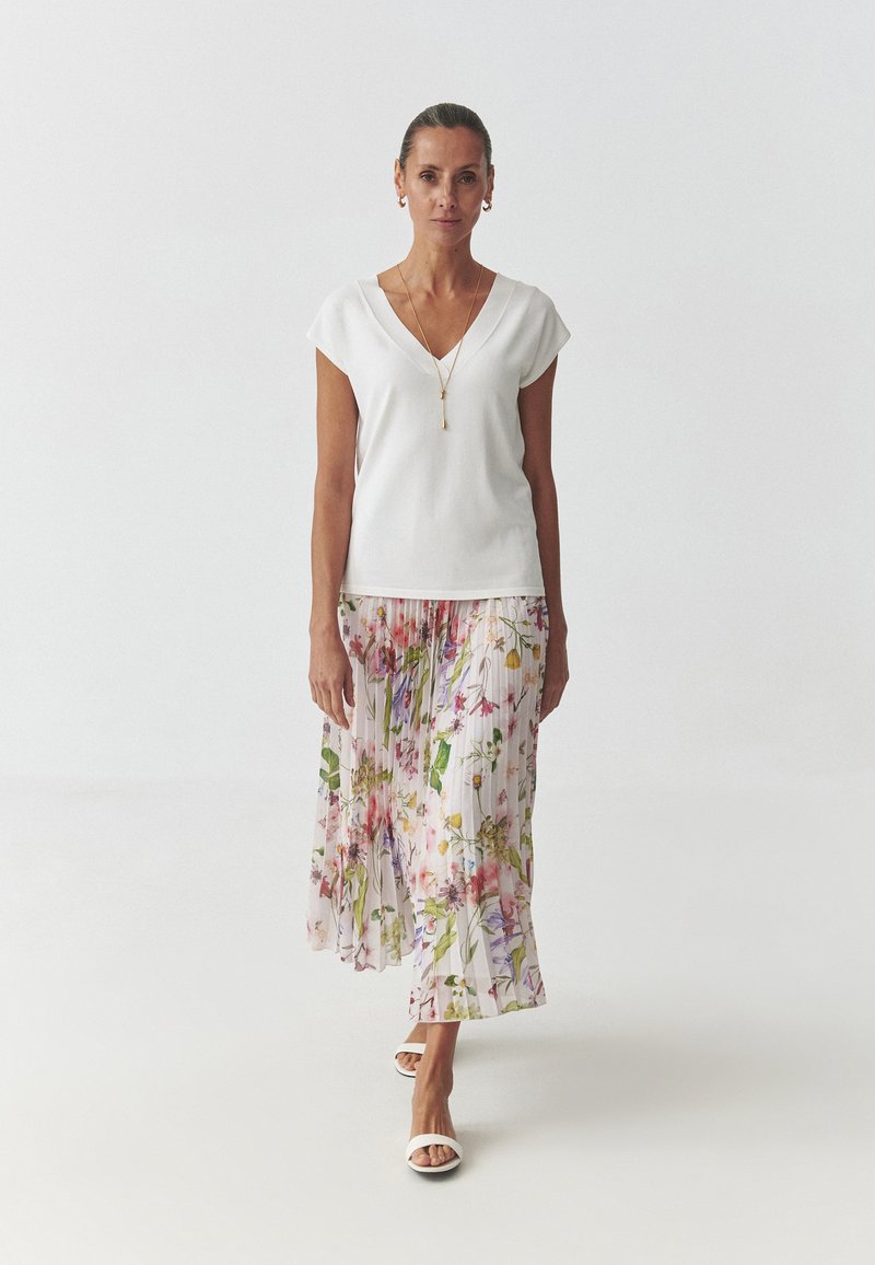 Top bianco con scollatura a V abbinato a una gonna fluida e plissettata con stampa floreale in rosa, giallo e verde. Modella che indossa sandali bianchi.