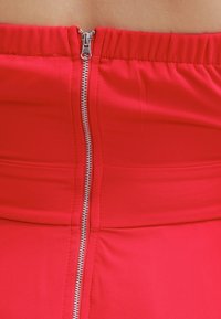 Rød nederdel med en glat stoftekstur, der har en synlig lynlås bagpå. Livremmen er elastisk for et tætsiddende udseende.