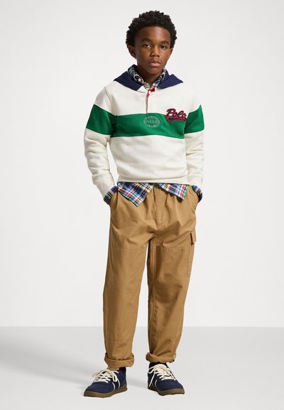 Sweatshirt com capuz em creme, verde e marinho com logotipo; camisa xadrez por baixo; calças cargo em caqui; ténis azuis com atacadores brancos.