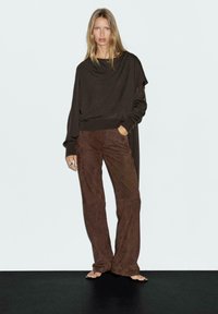 Brown knit top with draped neckline paired with brown suede wide-leg pants. Model stands with hands in pockets against a light background.