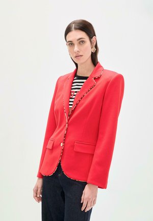 Blazer rojo con corte entallado, ribete dorado, dos bolsillos delanteros y cierre con un solo botón, llevado sobre una camisa a rayas y jeans oscuros.