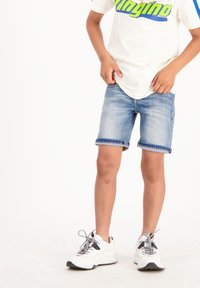 Ljusblå denimshorts med uppvikta ärmar, matchade med en vit t-shirt med grön och blå text, samt vita sportskor.