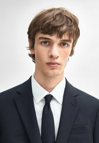 Jonge man met kort bruin haar, gekleed in een donker pak, witte shirt en een zwarte stropdas. Neutrale achtergrond, geconcentreerde uitdrukking.