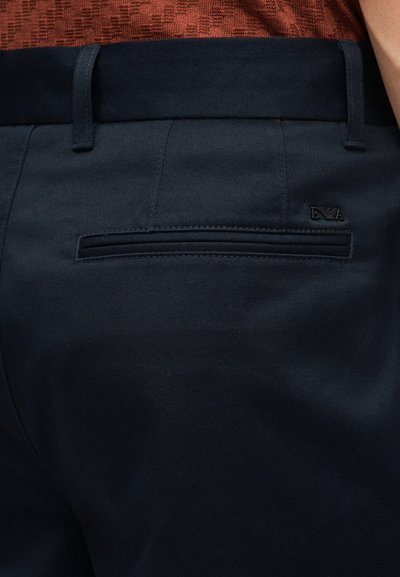 Κοντινό πλάνο σε ναυτικό μπλε παντελόνι ραμμένο στα μέτρα με οπίσθια τσέπη τύπου wel και μικρό μεταλλικό λογότυπο, φορεμένο με υφασμάτινο πουκάμισο σε σκουρόχρωμη σκουριά.